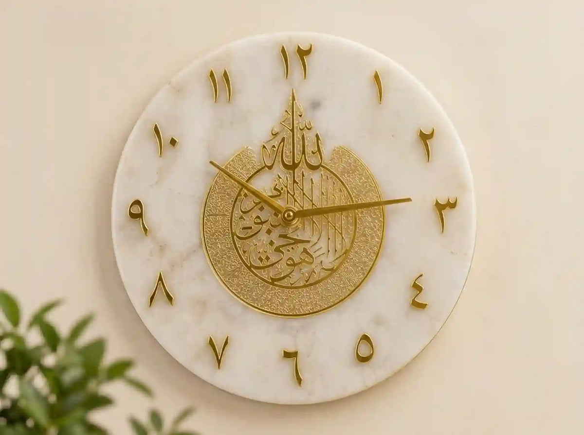 A round white marble wall clock with gold Arabic numerals and a central gold Islamic calligraphy design, mounted on a beige wall with a small green plant visible in the lower left corner.