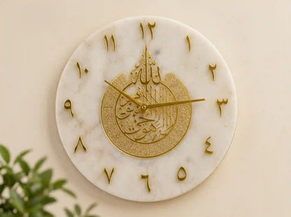A round white marble wall clock with gold Arabic numerals and a central gold Islamic calligraphy design, mounted on a beige wall with a small green plant visible in the lower left corner.