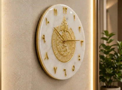 An oval wall clock with a white marble-look face and ornate gold details, mounted on a beige wall with a softly lit interior and a plant visible in the background.
