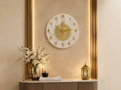 A round white marble wall clock with gold Roman numerals and gold hands, mounted on a beige wall with warm backlighting. Below the clock, a console table holds a vase with white blossoms, a candle, a book, and a gold lantern.
