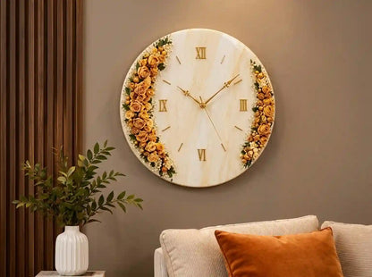 A round wall clock with a cream marble-look face, gold Roman numerals, and gold hands, decorated with yellow and orange roses around the left and right sides. The clock is mounted on a brown wall above a sofa with an orange pillow, next to a white vase with green leaves.